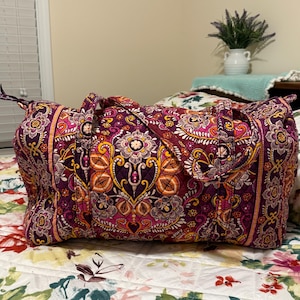 May include: A quilted duffel bag featuring a vibrant purple, pink, and orange floral paisley pattern. The bag has two fabric handles, a top zipper closure with a branded pull, and a rectangular structure with rounded corners and quilted stitching.