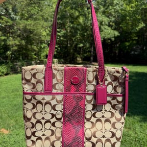 May include: A beige and brown monogram print tote bag featuring a vertical center panel with a bright pink snakeskin texture. The bag includes magenta leather trim, two matching shoulder straps, and a hanging leather tag with gold hardware.