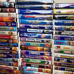 Puede incluir: Una gran colección de cintas VHS apiladas que presentan varias películas animadas y de acción real. Los lomos muestran títulos como Beauty and the Beast, Aladdin, The Brave Little Toaster, Monsters, Inc., Cinderella, Sleeping Beauty y Mulan.