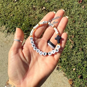 May include: A white beaded bracelet with the name "CHARLIE KIRK" spelled out in black letters. The bracelet includes a black cross charm and gold-coloured beads. The bracelet is held in a hand.