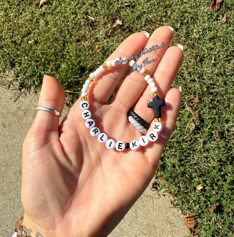 May include: A white beaded bracelet with the name "CHARLIE KIRK" spelled out in black letters. The bracelet includes a black cross charm and gold-coloured beads. The bracelet is held in a hand.