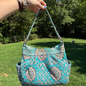 Könnte beinhalten: Eine gesteppte Stoffhandtasche mit einem Paisley-Muster in Türkis, Beige und Dunkelbraun. Die Tasche verfügt über einen einzelnen Schulterriemen, der mit silberfarbenen Metallringen befestigt ist, einen rechteckigen Boden und offene Seitentaschen.