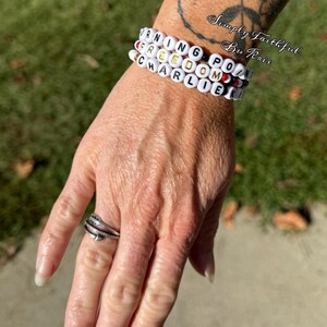 May include: Three white beaded bracelets spelling out "WARNING FREEDOM POINT CHARLIE" in black and gold letters. The bracelets are stacked on a wrist. A silver ring with a feather design is on a finger.