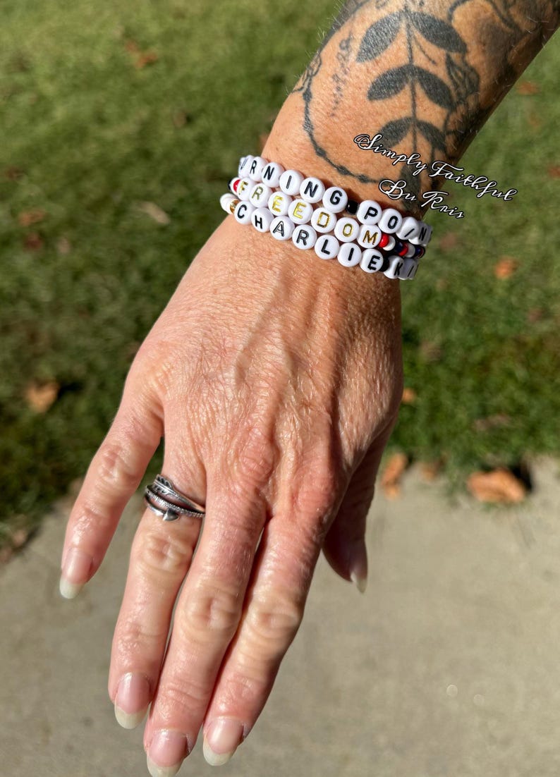 May include: Three white beaded bracelets spelling out "WARNING FREEDOM POINT CHARLIE" in black and gold letters. The bracelets are stacked on a wrist. A silver ring with a feather design is on a finger.