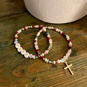 Può includere: Tre braccialetti di perline con un ciondolo a forma di croce dorata. Un braccialetto ha perline bianche con la parola "PRAY" scritta in lettere dorate. Gli altri due braccialetti hanno perline rosa, rosse e verdi.