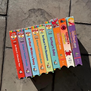 Puede incluir: Una colección de cintas VHS coloridas con series animadas para niños. Los títulos incluyen "Arthur's Chicken Pox", "Arthur's Teacher Trouble" y "Clifford the Big Red Dog". Las cintas están dispuestas sobre una superficie texturizada.