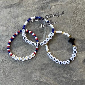 May include: Three beaded bracelets with white letter beads spelling "FREEDOM", "TURNING POINT", and "CHARLIE KIRK". The bracelets feature red, white, and blue beads, and gold-coloured beads. The bracelets are on a grey surface. The text "Simply Faithful By Kris" is in the background.