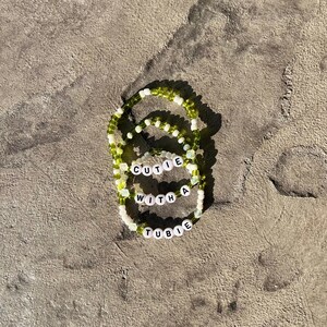 Op de afbeelding: Drie kralenarmbanden met groene en witte kralen. De armbanden spellen "CUTIE WITH A TUBIE".
