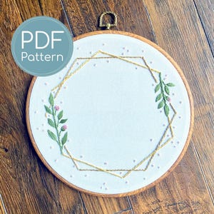 May include: A white fabric embroidery hoop with a geometric gold design and floral accents. The embroidery features green leaves and pink flower details. The text "PDF Pattern" is in a blue circle in the upper left corner.