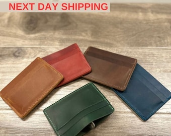 May include: Five leather card holders in different colors: brown, red, dark brown, blue, and green. The card holders are laid out on a wooden surface.