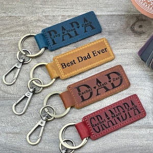 May include: Four leather keychains with metal rings and clasps. The keychains are personalized with the words "DADA", "Best Dad Ever", "DAD", and "GRANDPA" with names and hearts engraved below.