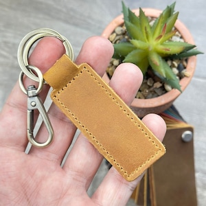 May include: A tan leather keychain with a stitched border and a silver key ring. The keychain is held in a hand.