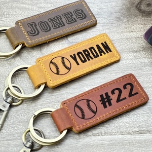 Könnte beinhalten: Drei Leder-Schlüsselanhänger mit Baseball-Motiven. Der obere Schlüsselanhänger ist braun und trägt die Aufschrift "JONES". Der mittlere Schlüsselanhänger ist beige und hat eine Baseball-Grafik und trägt die Aufschrift "YORDAN". Der untere Schlüsselanhänger ist braun und hat eine Baseball-Grafik und trägt die Aufschrift "#22".