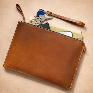 May include: A tan leather clutch bag with a zip closure, holding a smartphone, keys, and papers. The bag has a wrist strap and is made of smooth, textured leather. The keys have a blue key fob.