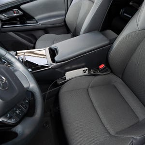 Op de afbeelding: Auto-interieur met een zwarte console met een ingebouwde telefoonhouder. Een zilveren smartphone is aangesloten op een oplaadkabel. Het stuur en de voorstoel zijn zichtbaar, samen met een rode veiligheidsgordelknop.