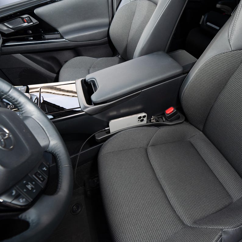 Op de afbeelding: Auto-interieur met een zwarte console met een ingebouwde telefoonhouder. Een zilveren smartphone is aangesloten op een oplaadkabel. Het stuur en de voorstoel zijn zichtbaar, samen met een rode veiligheidsgordelknop.