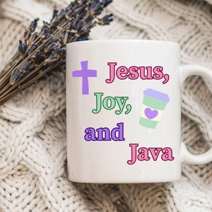 May include: White ceramic mug with the words "Jesus, Joy, and Java" in pink, green, and purple. A purple cross and a coffee cup with a heart are also on the mug. The mug has a curved handle.