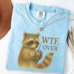 May include: Light blue t-shirt featuring a raccoon holding a walkie-talkie. The text "WTF, OVER" is printed in brown. The shirt is folded with a necklace and a hat visible in the background.