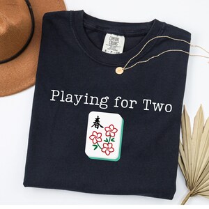 May include: Navy blue t-shirt with the text "Playing for Two" and a Mahjong tile graphic. The tile features red flower designs and Chinese characters. A gold necklace and a brown hat are also visible.