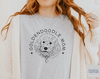 Puede incluir: Una camiseta gris con una ilustración en blanco y negro de un perro golden doodle con un collar con un dije en forma de corazón. El texto "Golden Doodle Mom" está escrito en un círculo alrededor del perro.