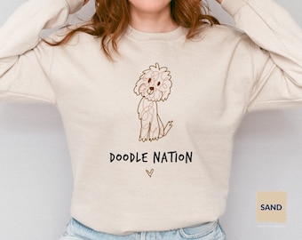 以下が含まれることがあります： ベージュのトレーナーに、犬の落書きと「Doodle Nation」という茶色の文字が書かれています。文字の下には小さなハートがあります。