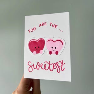 Könnte beinhalten: Eine weiße Grußkarte mit dem Text "You are the... Sweetest" in roter Schrift. Die Karte zeigt zwei herzförmige Charaktere, einen dunkelrosa und einen hellrosa, mit lächelnden Gesichtern.