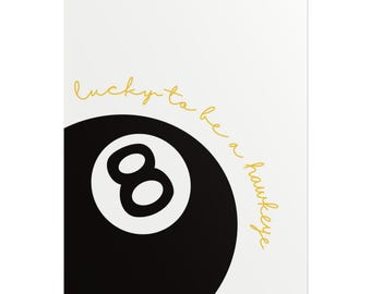 May include: A white background with a black eight ball and the number 8 in white. The text "lucky to be a hawkeye" is written in gold script.