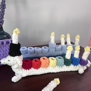 May include: A crocheted dachshund dog menorah. The dog is white with a black hat. The candle holders are in rainbow colours, and the candles are white with yellow tips. A detached candle is also present. The menorah is approximately 30 cm long.