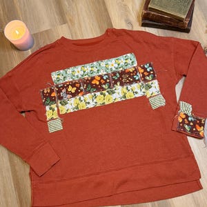 May include: A rust-colored long-sleeve sweatshirt featuring a central horizontal panel of patchwork fabric. The patchwork includes floral patterns in yellow and green, and a dark brown fabric with a butterfly print. Similar patches appear on the cuffs.