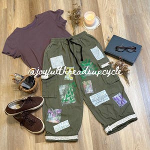 May include: An olive green pair of cargo pants with patches of various designs, a brown t-shirt, and brown sneakers are arranged on a wooden surface. The pants have a drawstring waist and lace trim at the ankles. The text "@joyfulthreadsupcycle" is visible.