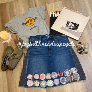 May include: A denim skirt with a patchwork design, a gray Hard Rock Cafe Nashville t-shirt, and a vintage album are displayed. The skirt has a dark wash with a decorative trim and colorful fabric circles. A pair of olive-green sneakers and a lit candle are also present.