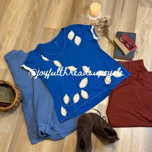 May include: A flat lay featuring a blue t-shirt with leaf-like embroidery, a blue sweater, and burgundy pants. Brown suede shoes and a small basket are also present. A lit candle, dried flowers, and books complete the scene.
