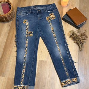 May include: Blue denim jeans with sunflower patchwork details. The jeans feature sunflower fabric strips down the sides and on the pockets, with rolled-up cuffs. The jeans are displayed on a wooden surface.