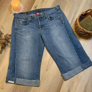 May include: Light blue denim capri jeans with a button fly and rolled cuffs. The jeans have a faded wash and classic five-pocket styling. A small logo is visible on the cuff. The jeans are folded on a wooden surface.