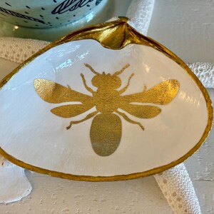 May include: A white seashell dish with a gold painted rim and a gold bee design in the center. The dish is on a white surface with a starfish and a decorative bowl in the background. The dish is likely used for jewelry or small items.
