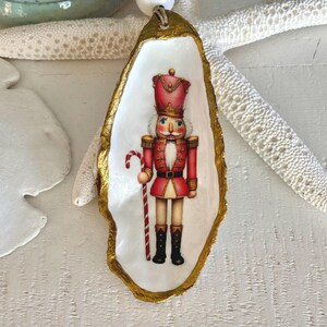 May include: A decorative oyster shell featuring a painted image of a nutcracker soldier in a red uniform with a candy cane. The shell has a gold-painted edge and is displayed on a white surface with a starfish.