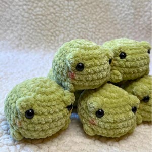 May include: A group of small, light green crocheted frog plushies made from soft yarn. Each frog features round black plastic eyes and small pink stitched blush marks on the cheeks. The frogs have a rounded, textured body shape.