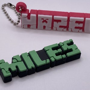 May include: Two personalized keychains with names "JAEL" and "MILES" in blocky 3D letters. The keychain with the name "JAEL" is red with white letters. The keychain with the name "MILES" is green with black letters.
