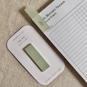 May include: A pack of green sticky notes labelled "Task Tabs" with the text "Vol 01 - Le Debut" and "Transparent 13x4.5cm - 30 Sheet". The sticky notes are on a beige fabric surface.