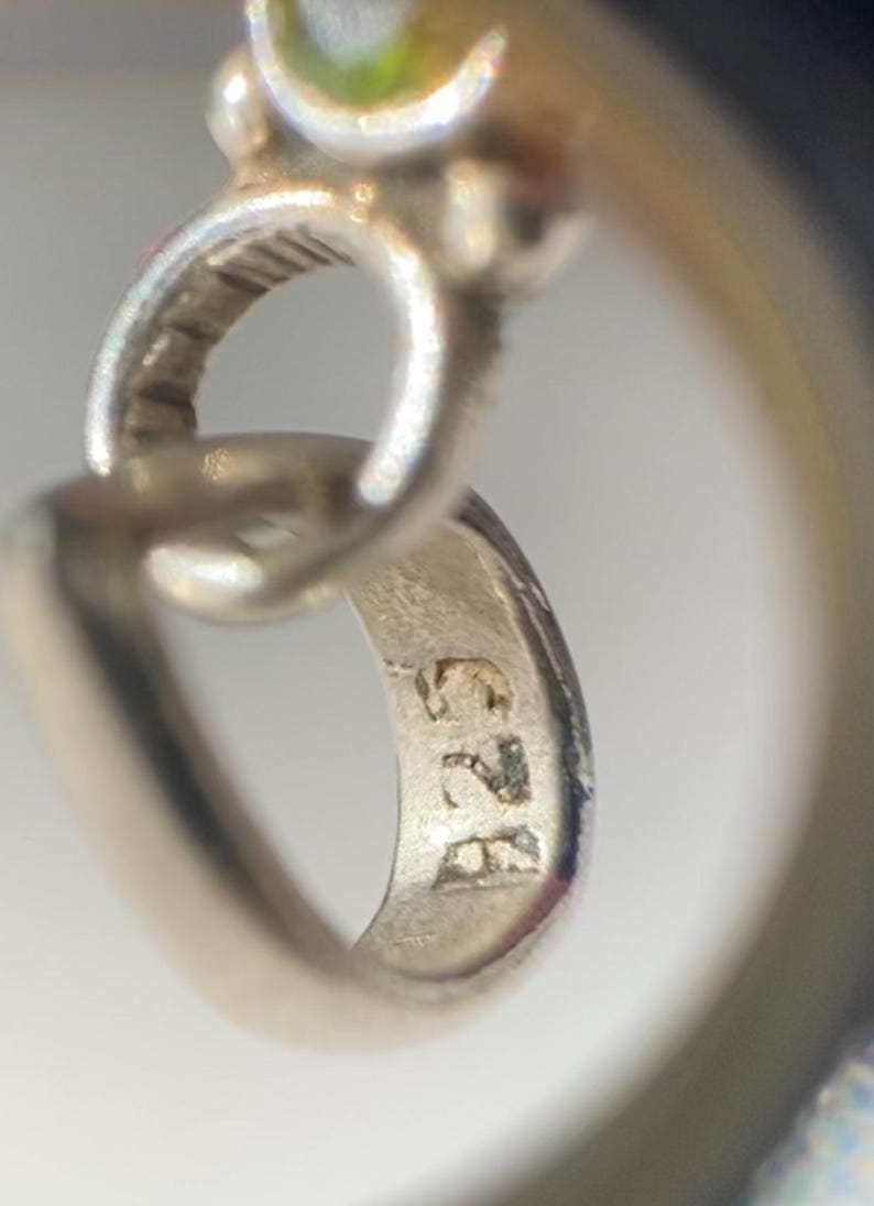 Peut inclure: Gros plan d'un pendentif en argent avec une pierre pr&eacute;cieuse verte. Le pendentif a une boucle pour l'attacher &agrave; une cha&icirc;ne et est marqu&eacute; du num&eacute;ro "925" au dos.
