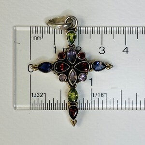 Peut inclure: Un pendentif en forme de croix en argent avec une vari&eacute;t&eacute; de pierres pr&eacute;cieuses color&eacute;es, notamment du rouge, du vert, du violet et du bleu. Le pendentif mesure environ 1 pouce de long.