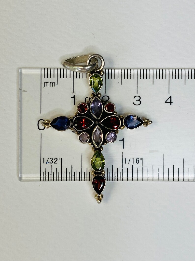 Peut inclure: Un pendentif en forme de croix en argent avec une vari&eacute;t&eacute; de pierres pr&eacute;cieuses color&eacute;es, notamment du rouge, du vert, du violet et du bleu. Le pendentif mesure environ 1 pouce de long.