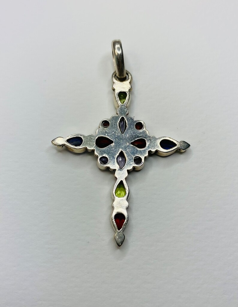 Peut inclure: Un pendentif en forme de croix en argent avec des pierres pr&eacute;cieuses color&eacute;es. La croix est d&eacute;cor&eacute;e de pierres pr&eacute;cieuses rouges, vertes, violettes et bleues.
