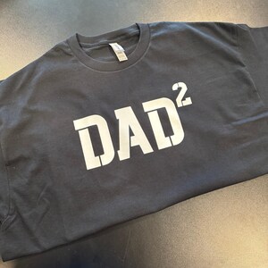 Op de afbeelding: Een zwart T-shirt met het woord "DAD" in witte letters, met een superscript "2" na de "D".