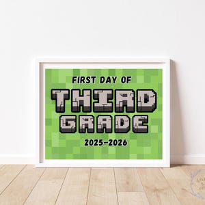 May include: A framed print with the text "FIRST DAY OF THIRD GRADE 2025-2026" in a pixelated font, set against a green pixelated background. The print is in a white frame and is displayed on a wooden floor.