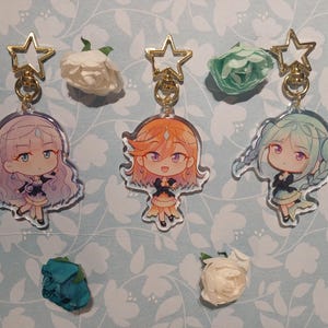 May include: Three clear acrylic keychains with gold star charms and cartoon character images. The characters are a girl with purple hair, a girl with orange hair, and a girl with green hair. Each character is wearing a different outfit.