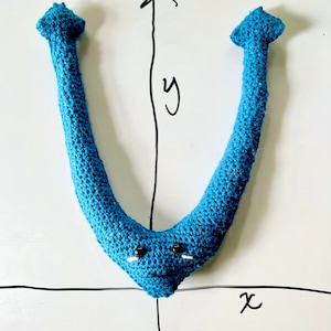 May include: A handmade, blue crochet V-shaped creature with black eyes and a small pink mouth. The creature is positioned on a white surface with a drawn coordinate plane, including the x and y axes.