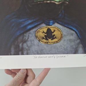 May include: Art print featuring a frog superhero with a blue cape and a frog emblem. The print includes the text "The Hero we need & deserve" and is numbered 1/10. The artwork has a vintage aesthetic.