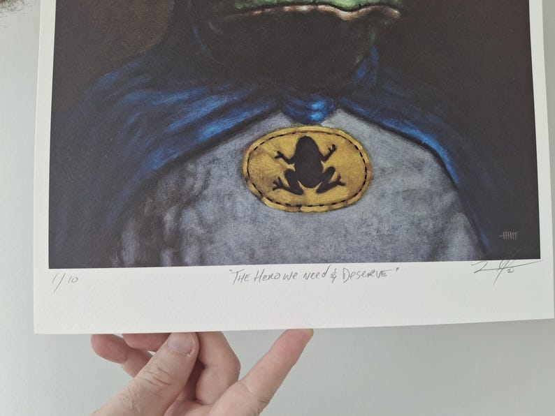 May include: Art print featuring a frog superhero with a blue cape and a frog emblem. The print includes the text "The Hero we need & deserve" and is numbered 1/10. The artwork has a vintage aesthetic.