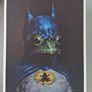 May include: A print featuring a frog dressed as Batman, with a blue cowl and cape. The frog has large, expressive eyes and a green face. A yellow oval emblem with a frog silhouette is on the chest. The text "The Hero we need & Deserve" is at the bottom.